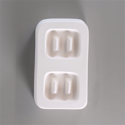 A rectangular white ceramic mold for fusing hot glass on a grey background. Two identical large square buttons, each with two long, thin posts to create holes for threading, have been carved into it with a space between them.