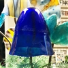 A fused glass mushroom on a copper tube stem displayed in front of other colorful glass flowers. The glass mushroom cap is dark blue and formed into a bell shape with a slightly waved bottom edge.