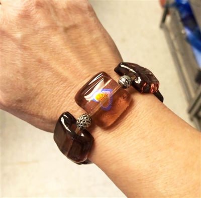 A close-up of a bracelet on a white womanâ€™s wrist. The bracelet is made of squares of lilac glass strung with metal beads between. The lilac square in the center also has a heart in it. The heart is blue outside, gold in the middle, and silver between.