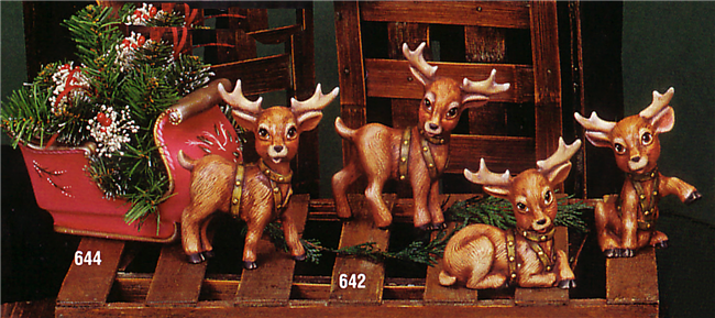 Four small ceramic reindeer in a line drawing a small red ceramic sleigh. Each reindeer is posed slightly differently, with the back two standing and front two sitting. The sleigh is filled with sprigs of evergreen.