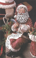A ceramic figure of a seated Santa Claus. He is dressed in the traditional red and white Santa clothing and has a long white beard. He holds a quill in his right hand and a long list of names in his left, and is looking kindly down at the list.