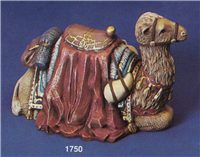 A ceramic figure of a seated camel on an indigo background. Its legs are tucked under it, and its fur is a light tan. It has a brown leather harness and ornate saddle on its hump. The saddle sits on red fabric that extends over the sides onto the ground.