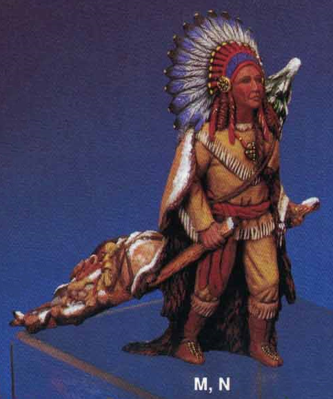 A cast ceramic figure of a Native American styled Santa. He wears leather clothing and a feather headdress and is carrying a travois with leather wrapped packages on it. He has an evergreen tree strapped to his back and wears a leather cape.