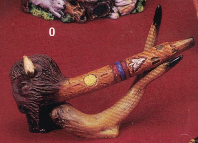 A cast ceramic piece made to look like a pipe with a buffaloâ€™s head as the bowl. The stem is textured like wood and decorated with geometric patterns. A separate cast ceramic stand made to look like an antler holds the pipe up at a diagonal.