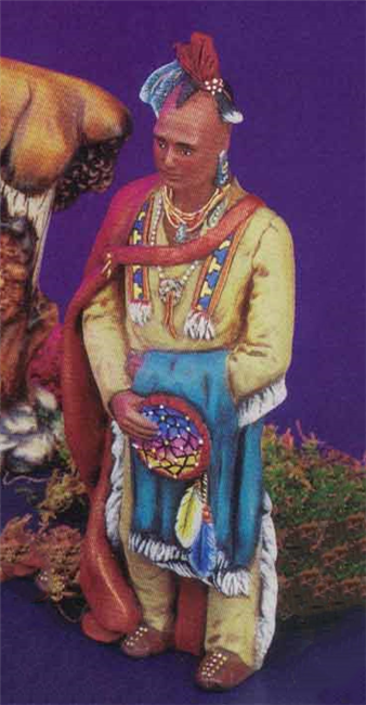 A cast ceramic figure of a Native American-styled wise man. His hair is a Mohawk decorated with feathers. He wears a hide jacket and pants with fringe, and has a red cape draped over his right shoulder. He holds a blue blanket and dream catcher.