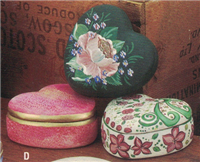 Three cast ceramic boxes. One is resting atop the other two, and all are heart-shaped. The bottom left is pink with gold trim. The bottom right is white with painted pink and green floral designs. The top is green with an intricate flower painted on top.