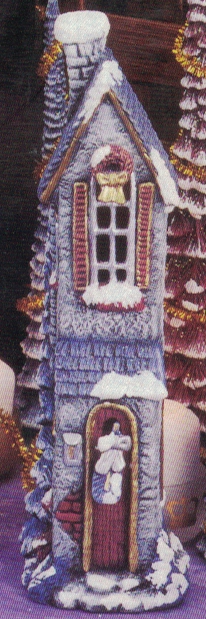 A cast ceramic figure of a tall, thin house. It has a tall evergreen tree behind it, and both the tree and house are capped in snow. The house has a chimney, a small cross on the door, and a wreath atop the second-story window.