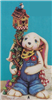 A cast ceramic figure of a long-eared rabbit in overalls. It stands, holding a post wrapped with vines in its right hand. The post is topped with a birdhouse and a small bird sitting atop it. The rabbit also has a scarf tied around its neck.