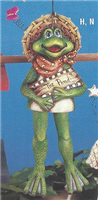 A cast ceramic figure of a green frog. It is hung from a branch and its legs are attached in such a way that they dangle from its body and move as it moves. The frog wears a straw hat and scarf and holds a sign that says The Pond.