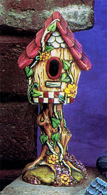 A cast ceramic figure of a birdhouse atop a tree truck. There are vines and flowers growing up the trunk onto the birdhouse. The house is yellow with a red roof and red-and-white checkered trim, and its opening is oval.