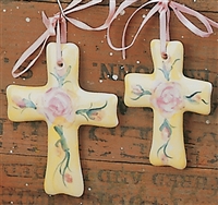 Two flat cross ornaments made of cast ceramic. The left is slightly longer. Each has a yellow base with a green and pink floral design painted on it. They are hung with thin pink ribbons.
