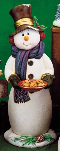 A large cast ceramic snowman made of multiple parts. He has a top hat, earmuffs, and long scarf. He wears mittens and his arms are outstretched in front of him holding a plate with real cookies on it. The base looks like a pile of snow with greenery in it