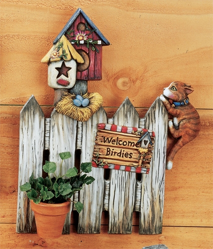 A cast ceramic piece of a section of picket fence. There are two birdhouses on posts and a small nest of eggs below them. A cat hangs off the right side, as does a sign reading Welcome Birdies. There is a small pot attached at the bottom.