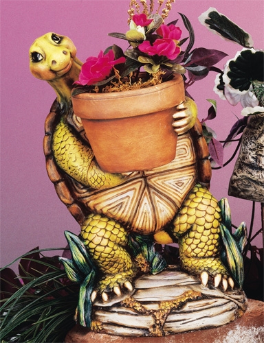 A cast ceramic figure of a large turtle standing atop a rock, holding a flower pot in its outstretched arms. Its legs are bent slightly and it looks forward. The rock has greenery around it, sprouting up around the legs of the turtle.