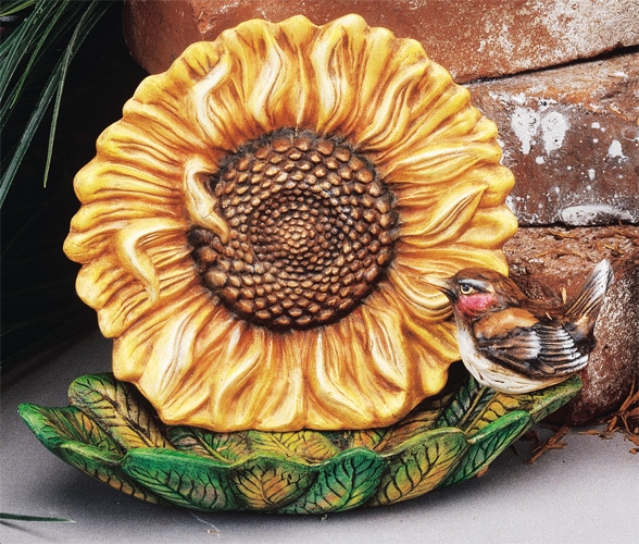 A birdfeeder made of several pieces of cast ceramic. The dish is shaped like a bundle of leaves. Behind it is a cupped disk in the shape of a large sunflower. A small cast ceramic bird, painted like a house sparrow, sits on the edge of the dish.