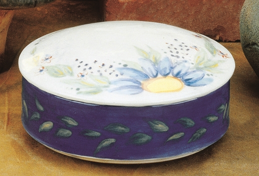 A round cast ceramic box with a round cast ceramic lid. The box is painted dark blue with green leaves around it. The lid is white with a floral design painted around its circumference.