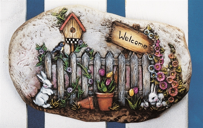 A cast ceramic stone slab that is oval in shape and flat on either side. It shows a picket fence surrounded with flowers, flower pots, and rabbits. There is a birdhouse on a post behind the fence on the left, and a sign reading Welcome on the right.
