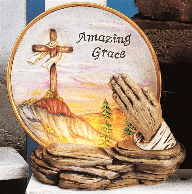 A round cast ceramic plate placed into a base of cast ceramic made to look like rock. The plate is embossed with a cross atop a rocky mountainside. The words Amazing Grace are carved in the sky beside it. A pair of praying hands is also on the rock base.