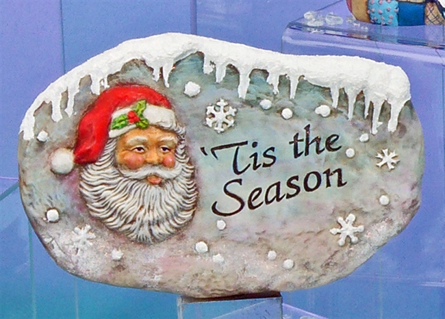 A cast ceramic stone slab that is oval in shape and flat on either side. It shows a traditionally styled Santa face on the left with the words â€˜Tis the Season carved on the right. There are snowflakes scattered around, and icicles hang from the top.