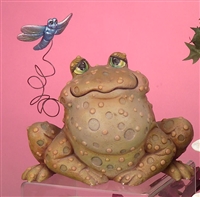 A large cast ceramic figure of a seated toad. It looks forward, eyes half closed. Its body is covered in small spots and bumps. A cast ceramic dragonfly is attached to it with a spiraling wire, allowing it to bounce.