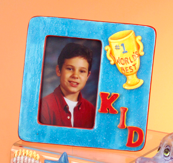 A cast ceramic picture frame with smaller cast ceramic pieces placed on it as decoration. The photo, of a young boy with dark hair, is on the left. A casting of a trophy reading Worldâ€™s Best is placed above three letters spelling KID below.