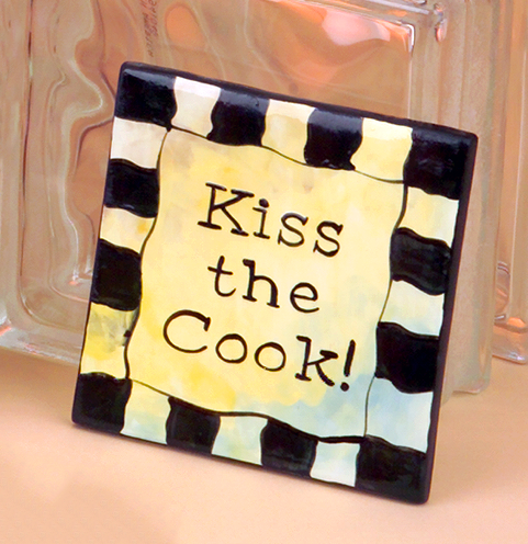 A square tile made of cast ceramic. It is glazed off-white and has been painted with an outer border of black and white checkerboard. The words Kiss the Cook! are painted in the center to resemble typewriter font.