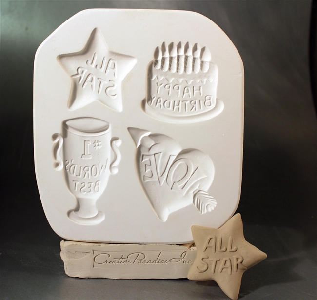 A white plaster mold for pressing moist clay into specific shapes. Four celebratory designs are carved into it. There is a cake with candles reading Happy Birthday, a star reading All Star, a trophy with no. 1 World Best, and a heart and arrow with Love