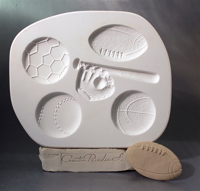 A white plaster mold for pressing moist clay into specific shapes. Five sport-themed shapes are carved into it. There is a football, baseball, soccer ball, basketball, and a baseball bat and glove joined together in a single cavity.