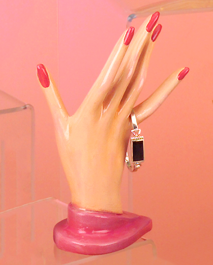 A cast ceramic figure of a feminine hand. The fingers are outstretched, each with a long painted nail. The hand rests atop a simple base. A metal bracelet dangles from the pinky finger.