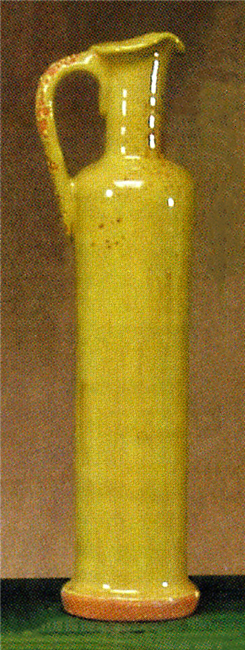 A tall, thin decanter made of cast ceramic for holding and pouring oil. It tapers into a thin neck at the top which then flares out again for pouring. The handle joins the top of the neck to the body. It is glazed with a light olive green color.