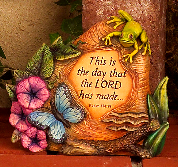 A cast ceramic figure of a rock surrounded by flowers, leaves, and a branch. A butterfly rests at the base of the rock and a frog is perched atop it. The center of the rock is flat and carved with the words of Psalm 118 verse 24.