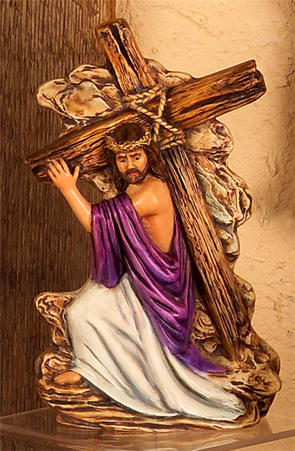 A ceramic figure of Jesus, kneeling in front of a wall of stone, carrying a wooden cross bound with rope over his right shoulder. He wears a crown of thorns and looks down. His robe slips from his shoulder.