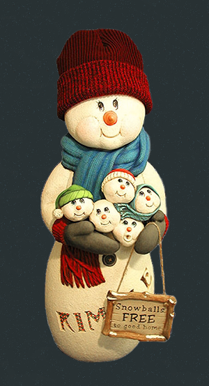A large cast ceramic snowman made of multiple parts. He has a non-ceramic knit cap on his head. He has a long scarf and mittens, which hold five snowballs with small snowman faces. He has a sign reading Snowballs free to good home hanging on his arm.