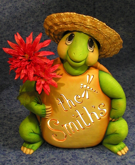 A large cast ceramic figure of a seated green turtle. It looks up happily. It holds real flowers in its right hand and has a real straw hat placed on its head. The words the Smiths and a small dragonfly are carved into its stomach with a light inside.