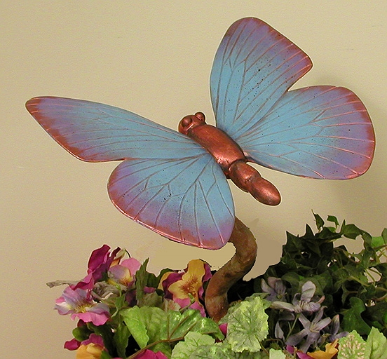 A large cast ceramic butterfly atop a curving cast ceramic stem. Its wings are outstretched and painted in a red to blue gradient. Its simplified body is bronze. The stem is placed into a pot with greenery.