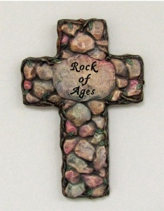 A flat cross of cast ceramic textured and painted to look as if assembled from many small stones stuck together. A large stone in the very center has the words Rock of Ages carved into it.