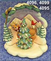 A scene made of multiple small cast ceramic figures. A deer looks around a decorated Christmas tree. It stands atop a base that looks like piled snow with a background decorated like a snowy landscape framed by trees and a pine branch behind it.