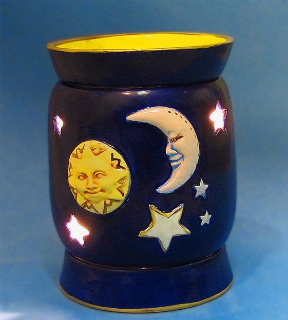 A three-piece cast ceramic wax melt burner. The circular top dish and bottom base are the same size and both glazed with dark blue. The cylindrical body is glazed the same color and shows a sun, moon, and stars. It has star-shaped holes to let light out.
