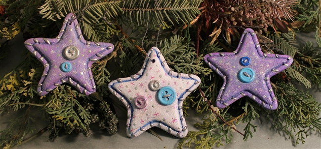 Three flat ornaments made of cast ceramic. They are all five pointed stars and shaped and textured to look as if made from fabric. They all have a visible seam with threads and each have a different configuration of small buttons stitched in the center.