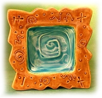 A picture of a square dish made from ceramic. It has irregularly wavy edges. The center depression, also square, is carved with a square swirl design and glazed teal. The outer rim is stamped and carved with a variety of geometric designs and glazed gold.