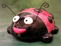A ladybug figure made of ceramic. Its body is round and hollow. It has two large round eyes, a smiling mouth, and four small balls as legs. It also has two curled wires as antenna. It is glazed with red and black.