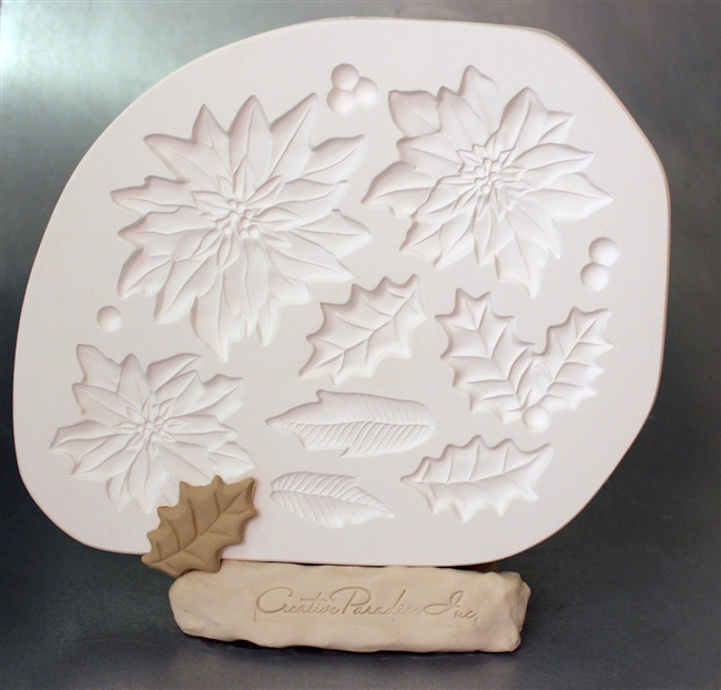 A white plaster mold for pressing moist clay into specific shapes.  Three separate poinsettia flowers of three sizes, two separate holly leaves, a holly bunch with two leaves, two separate lined leaves, and 3 separate clusters of berries are carved in it.