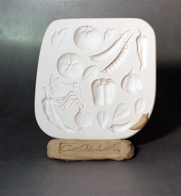 A white plaster mold for pressing moist clay into specific shapes. A variety of vegetable and leaf shapes are carved into it. There are two tomatoes, two green beans, three different chili peppers, a bell pepper, a garlic bulb, and a sprouting onion.