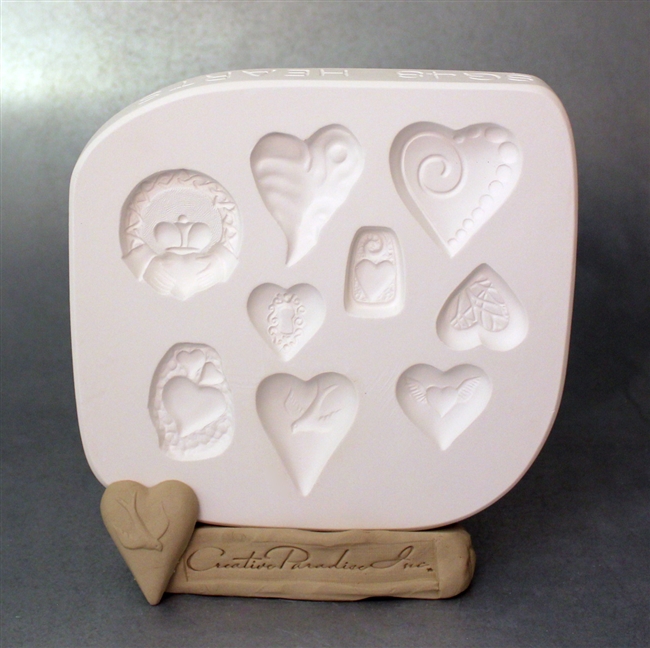 A white plaster mold for pressing moist clay into specific shapes. Nine heart themed shapes are carved into it. There are six different designs of heart, one heart within a rectangular tag, three hearts grouped together, and a traditional Celtic Claddagh.