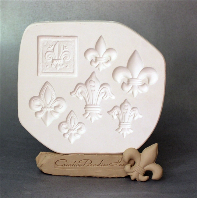 A white plaster mold for pressing moist clay into specific shapes. Seven fleur-de-lis shapes are carved into it. One is embossed within a square, while the others stand alone. They are in a variety of styles and sizes.