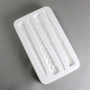 A white plaster mold for pressing moist clay into specific shapes. Four long, tapered handle shapes are carved into it. One has a texture of leaves, one is textured like wood, one has a braided texture, and one is textured with many clustered dots.