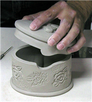 A hand lifting the lid of a finished but unfired ceramic dish. It is oval in shape, and the lid has a notch. The top of the box has a cross added on top and the sides are decorated with stamped floral designs.