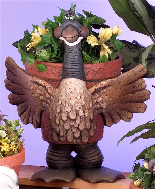 A large cast ceramic pot made of several pieces, shaped like a cartoonish goose. Its body is the pot, textured with false cracks around the rim. Its long neck comes up above the rim, and its legs and feet support the bottom. Its wings wrap around the pot.