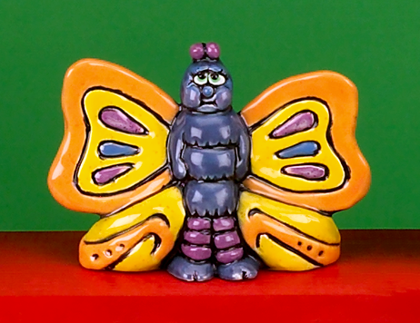 A cast ceramic figure of a butterfly with a more human like body. It stands upright on two feet, arms at its side. It looks forward with a look of reluctance in its large eyes and two round antennae on its head. Its large wings are stretched out.
