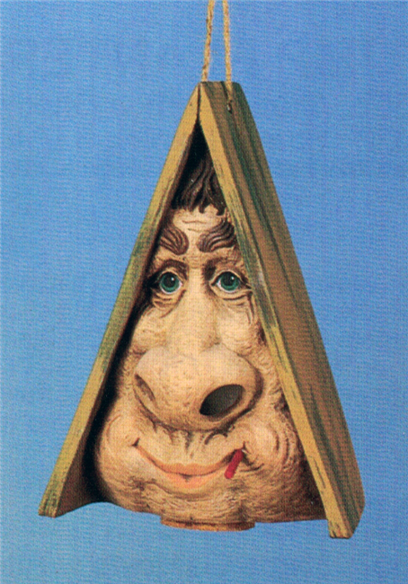 A cast ceramic figure of a cartoonish face hidden under a sloping A Line roof of wooden planks. The face has a very large nose and pursed, smiling lips. A stick protrudes from the lips, and the nostrils are carved out revealing a hollow interior.
